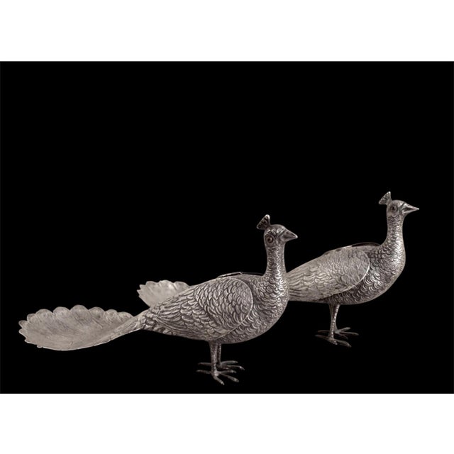 A pair of decorative silver peacock figurines with finely textured feather details and fan-shaped tail trays. Integrate...