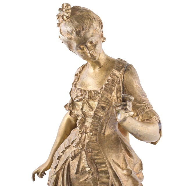 A French late 19th century figural bronze of a maiden in an 18th century ball gown. She is standing atop a circular...