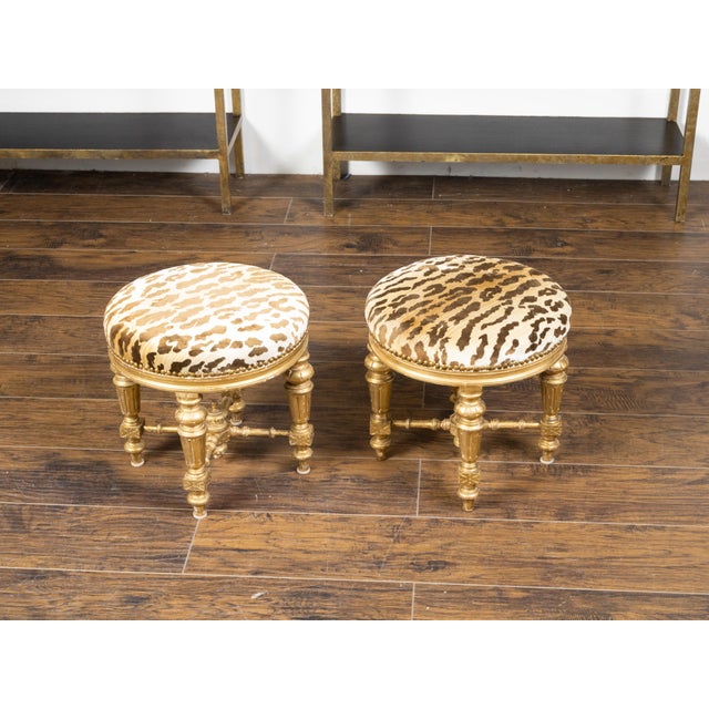 A pair of French giltwood stools from the 19th century, with fluted legs, cross stretchers and leopard style upholstery....