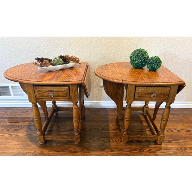 A handsome matched pair of vintage Riverside Furniture drop-leaf end tables featuring warm oak construction, beautifully...