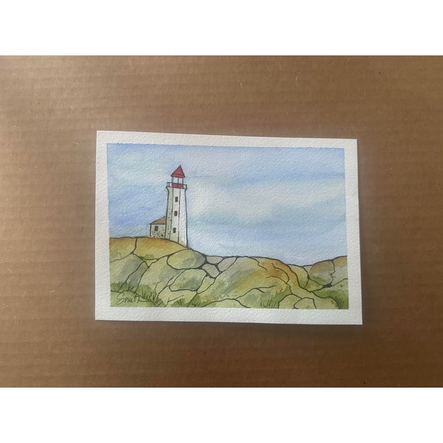 Original watercolor painting featuring a solitary lighthouse perched atop a rocky coastal bluff. Soft washes of blue sky...
