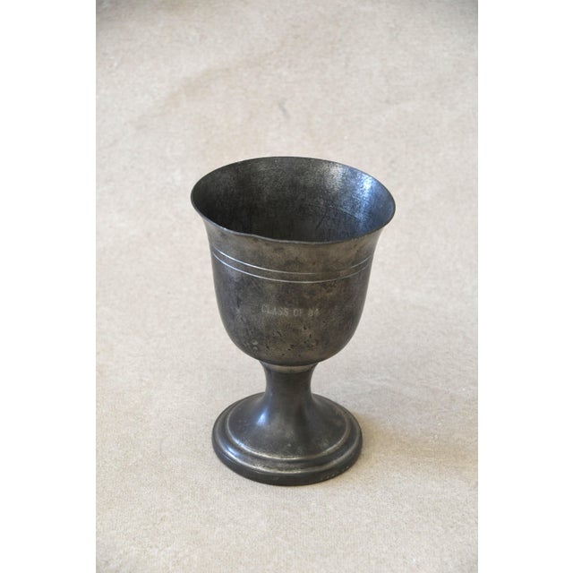 A footed pewter goblet engraved "CLASS OF 84," with banded ring detail below the rim. Warm, aged pewter finish with light...
