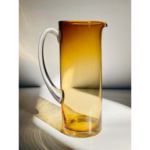 A transparent handmade glass pitcher in a beautiful rich amber glass shading from a darker hue at the top to a pale topaz...