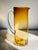 A transparent handmade glass pitcher in a beautiful rich amber glass shading from a darker hue at the top to a pale topaz...