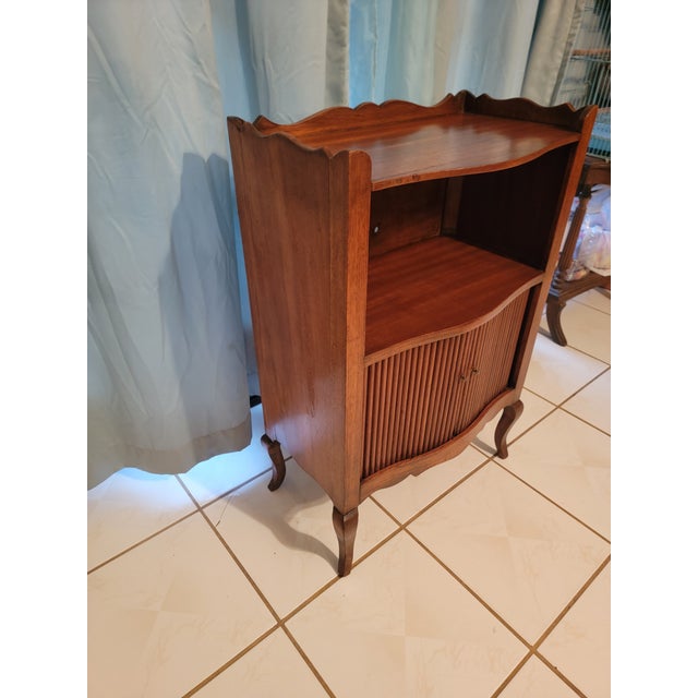 Antique wooden cabinet featuring a scalloped top, open shelf, and two tambour doors with storage inside. Circa early 20th...