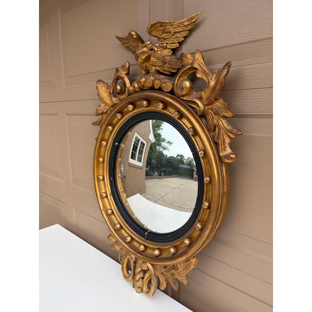 An antique Federal or Regency style giltwood convex mirror crowned with a carved eagle, its wings outstretched above a...