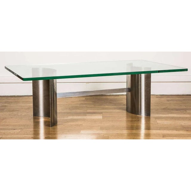 Rectangular coffee table with a stainless steel base and a thick glass top. The base is formed of two curved steel blades...