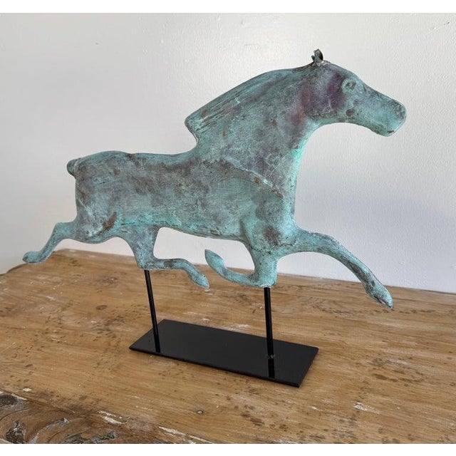 This sculptural piece is a vintage copper horse weather vane, artfully displayed on a modern black metal stand. The figure...
