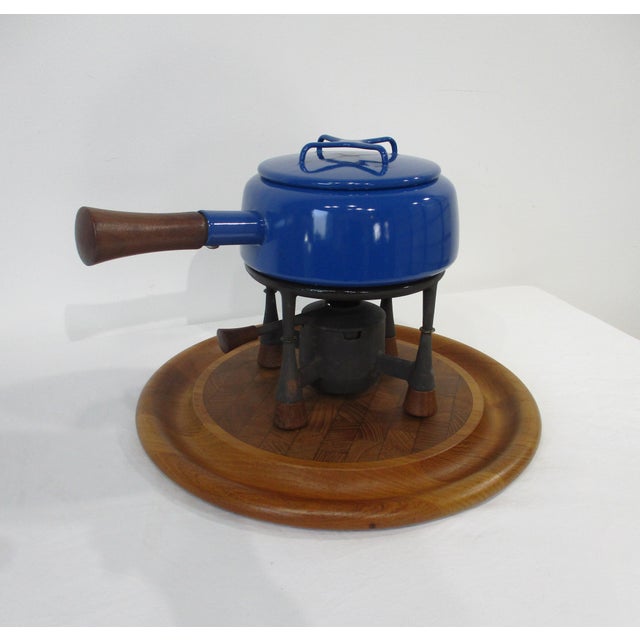 A three piece cooking set including a bright blue pot with lid , iron and teak pot holder sitting on a teakwood tray ....