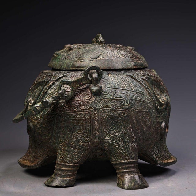 This is a cast bronze ritual vessel, featuring a robust, faceted body with a domed lid and a prominent arched handle. The...