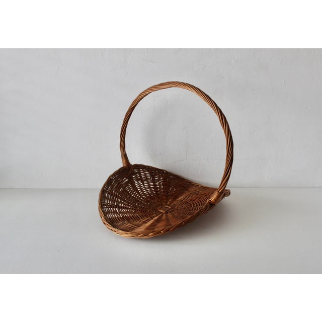 Vintage wicker catchall basket featuring a broad, shallow woven base and a tall arched handle. French-style, oval-shaped...