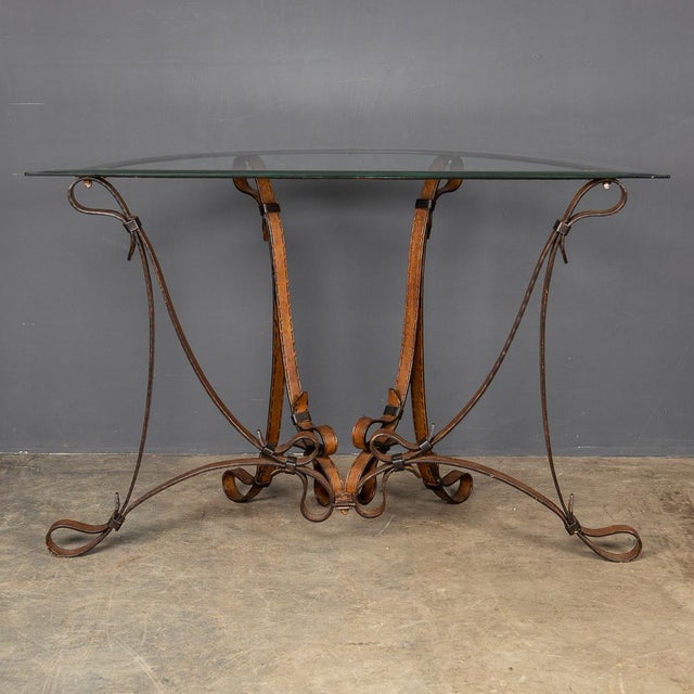 A sturdy cold painted wrought iron console, with stitched strap and buckle detail and a bevelled glass top, made in the...