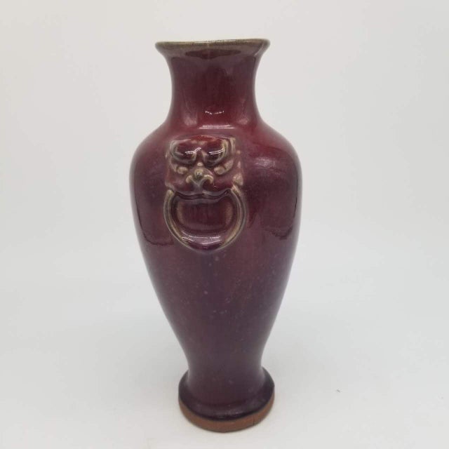 This is a vintage deep red ceramic vase featuring two sculpted lion-head handles on each side of the body. The lion masks...