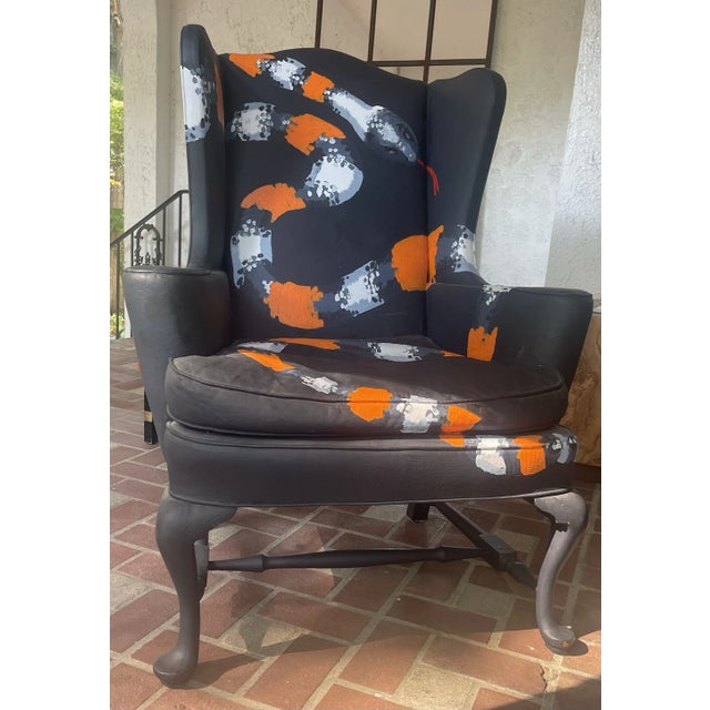 A vintage Queen Anne style wing chair painted black with painted upholstery with a painted orange and black snake that...