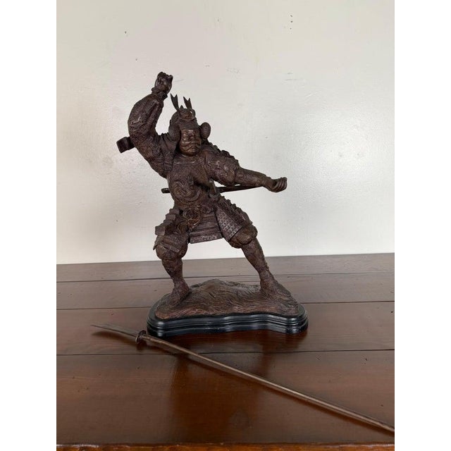 A dramatic Japanese bronze sculpture depicting a powerful samurai warrior captured mid-motion in a dynamic battle stance....