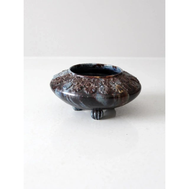 An antique three-footed bowl vase featuring an ornate cast floral design, a blue and brown painted glaze, and three...