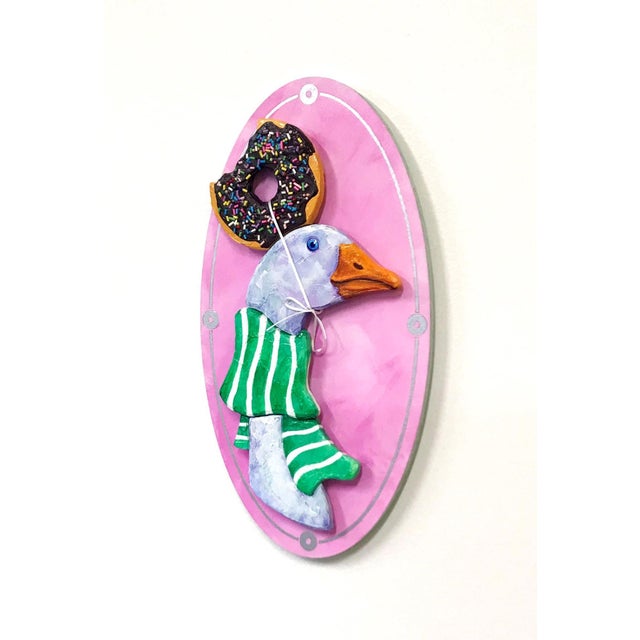 This artwork features a whimsical depiction of a goose adorned with a green striped scarf and a donut perched atop its...