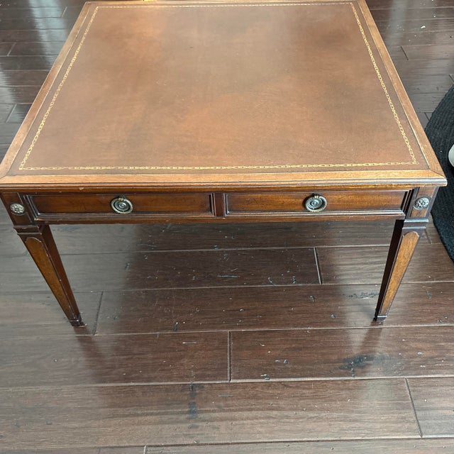 This vintage Baker Furniture coffee table features a brown leather-inset top accented with elegant gold tooling, classic...