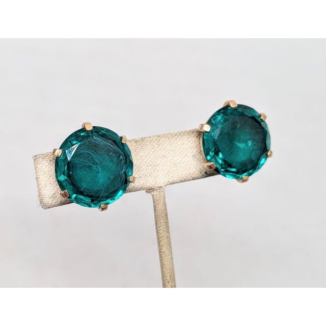 Early to mid 1950s round goldtone unfoiled open backed flawed faux-emerald clip earrings. Marked crown "TRIFARI" on the...
