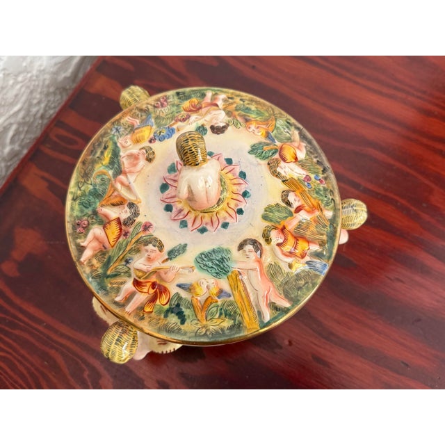 This round porcelain Italian Capodimonte lidded bowl is covered in raised relief cherubs at play among trees and flowers....