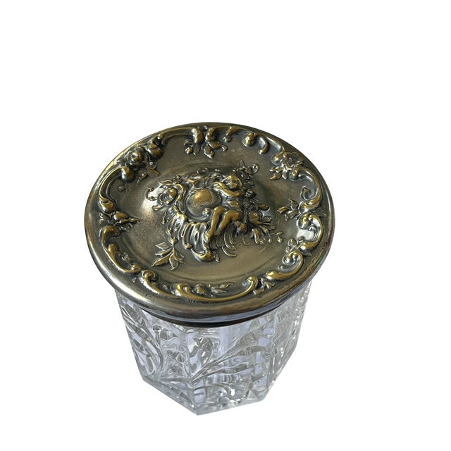 A beautifully detailed vintage jar featuring a substantial molded glass body paired with an ornate metal lid adorned with...