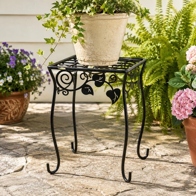 A charming vintage black metal garden plant stand featuring an open grid top, scrolling vine motif, stylized leaf details,...