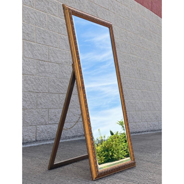 An elegant vintage full-length floor mirror featuring an ornate giltwood frame with carved foliate detailing and a warm,...