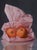 In "Papered in Pink," two vibrant oranges rest atop crumpled pink tissue, their glossy surfaces reflecting subtle light....