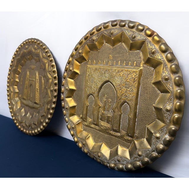 An eye-catching pair of vintage Moroccan repoussé brass wall chargers featuring intricate architectural scenes inspired by...