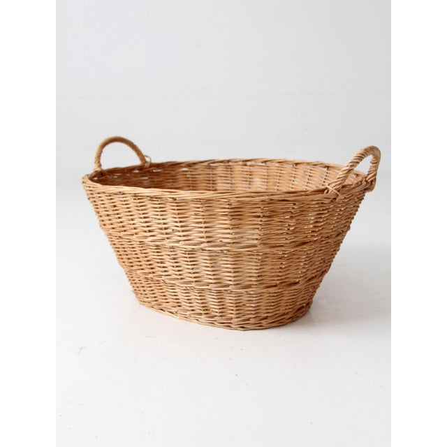 An antique wicker laundry basket with a broad oval shape, softly tapered sides, and two gracefully wrapped handles. The...