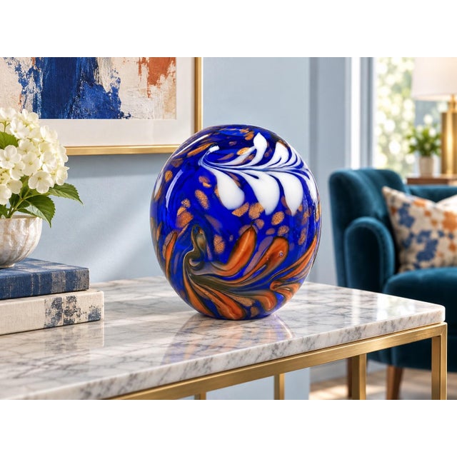 A striking art glass vase featuring a rich cobalt blue body decorated with flowing white, terracotta, and dark olive...