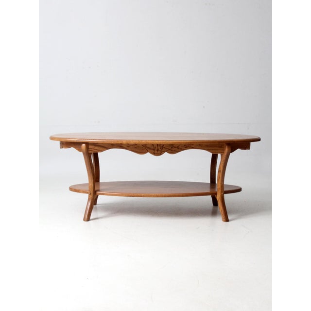 This vintage oak coffee table features curved legs and a carved wheat sheaf apron with an oval top and lower shelf. The...