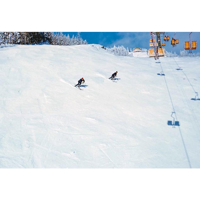Downhill Racers by Toni Frissell Two skiers on a piste go under a ski lift at the Stowe Mountain resort, Vermont, USA,...
