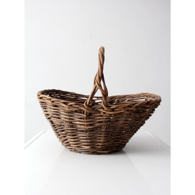 A vintage wicker gathering basket with a softly tapered silhouette and twisted overhead handle, woven in warm natural...