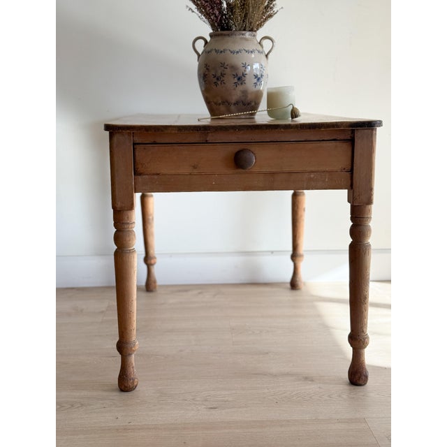 A square-cornered pine table on ring-turned legs, with a single working drawer closing on a dark turned knob. The top has...