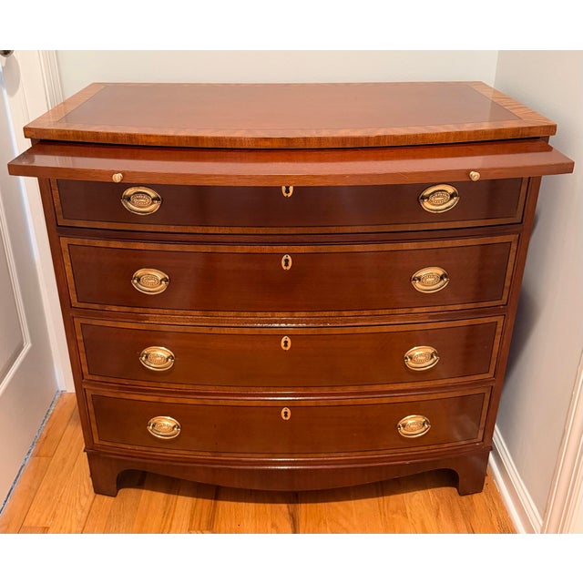 Lovely bowfront banded mahogany bachelors chest with 4 drawers and pull out shelf. Made by Baker Furniture Company, this...