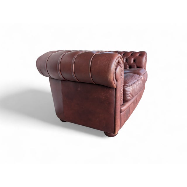 Chesterfield two-seater leather sofa in reddish brown/burgundy. The sofa shows signs of age, with scratches and wear on...
