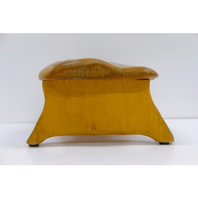 A low, sculptural footstool with a hand-shaped, live-edge wood top in a warm honey tone, showing natural grain variation...