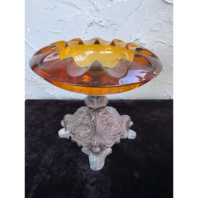 This elegant antique compote features a thick amber glass bowl with a sculptural scalloped rim, resting on an ornate cast...