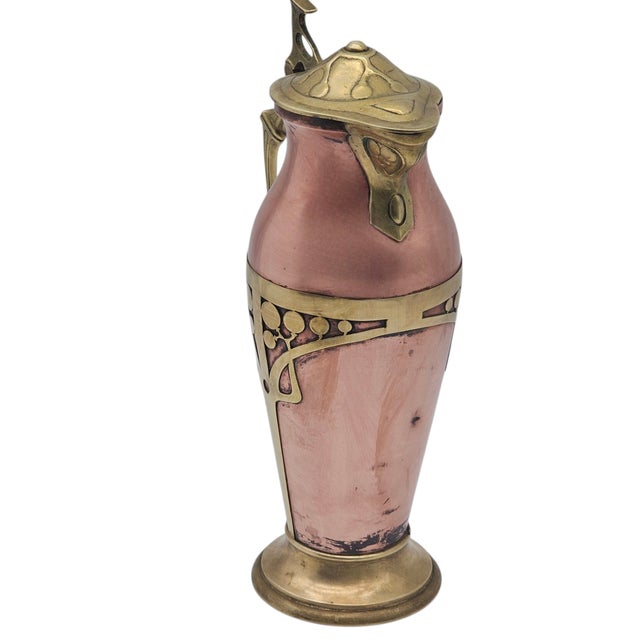 Here is a vessel of copper and brass, formed in the language of Art Nouveau. Its curves are softened by time, yet the...