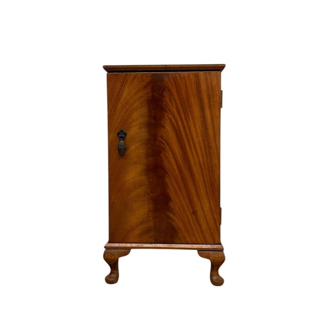 This bedside cabinet features a compact rectangular form with a richly figured veneer on the front door, displaying a...
