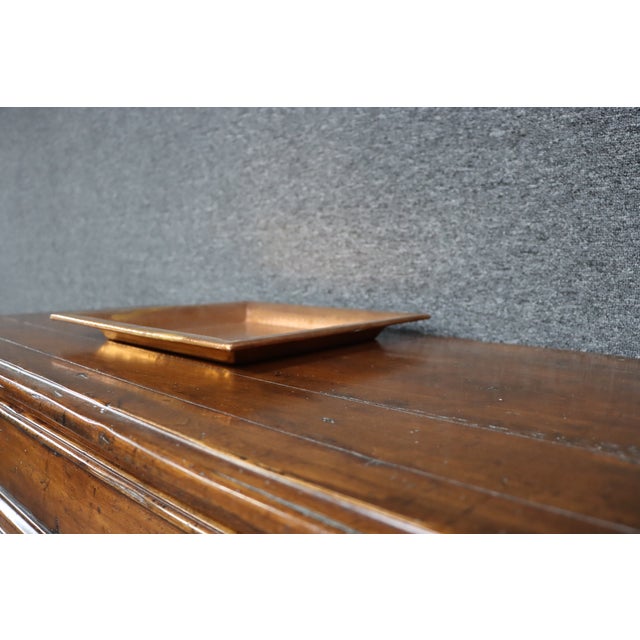 A hand-hammered copper tray featuring a clean square form with a wide angled gallery rim and an all-over dimpled finish....