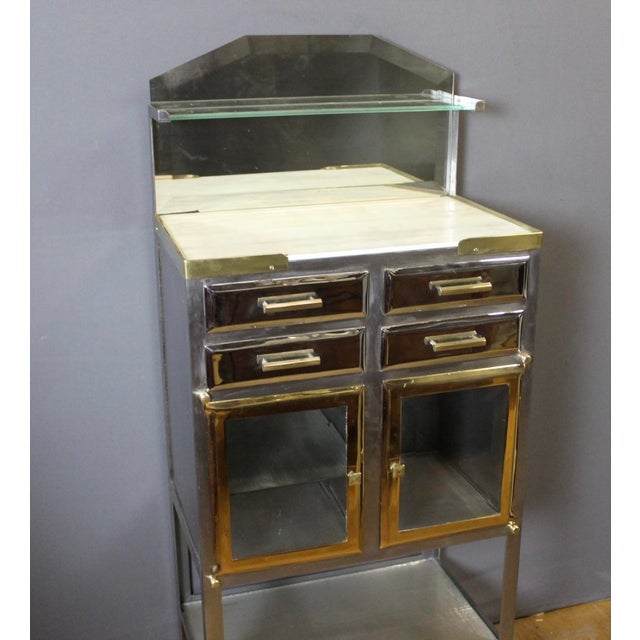 A dentist's cabinet in brushed, chromed, and gilded metal, featuring four drawers and two doors. The shelves are glass,...