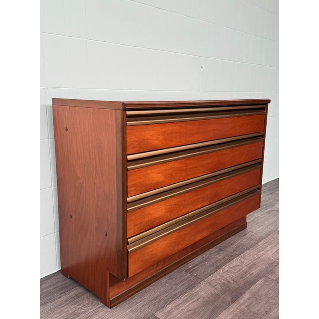 A 1970s-era wood veneer chest of drawers. The minimalist design features warm walnut veneer tones and is embellished with...