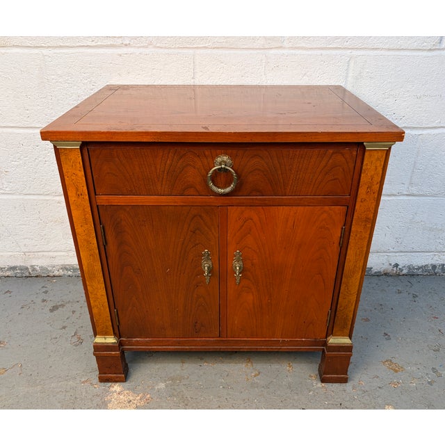 An elegant French Empire–style nightstand crafted in rich cherry with striking burled wood accents by Baker Furniture....