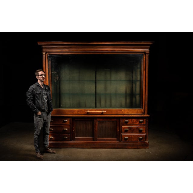 Monumental solid wood mercantile display cabinet featuring a full-width counter-balanced lift-front glass door, a design...