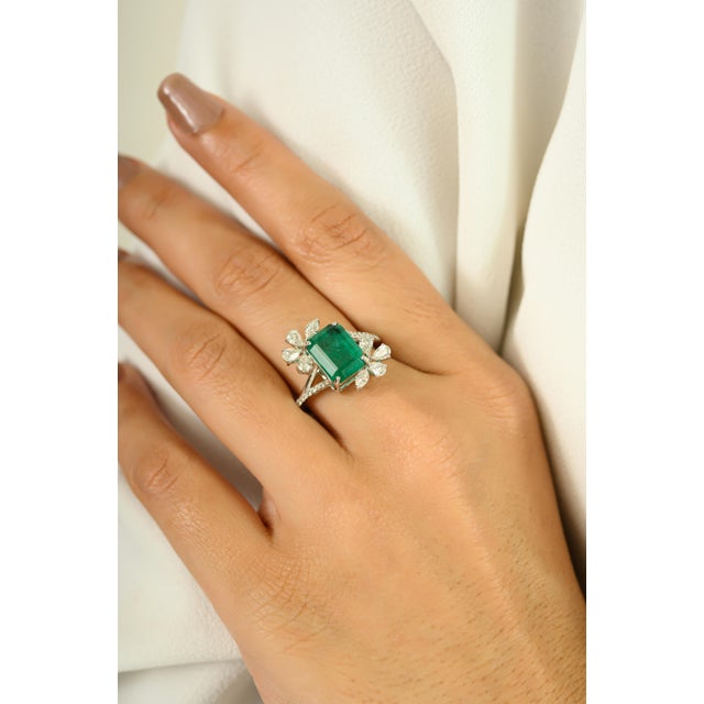 This dazzling ring showcases a vivid 3.56-carat emerald-cut emerald, beautifully framed by a delicate floral-inspired...