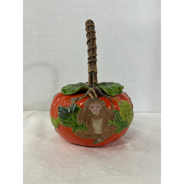 Small tomato shaped ceramic teapot with removable leaf shaped lid, monkey decoration, and woven handle.