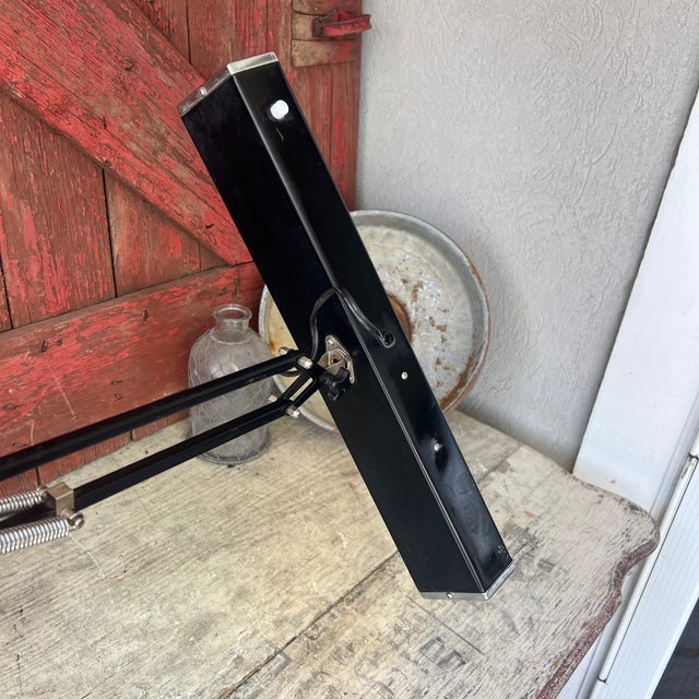This vintage desk lamp features a sleek black metal frame with spring-loaded articulating arms and a fluorescent light....