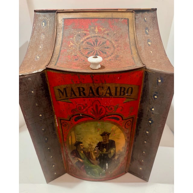 Striking antique tin tea caddy featuring bold MARACAIBO branding and a richly colored scenic figural panel on the front....